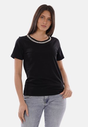 Femme portant un haut noir à manches courtes avec un col décoratif, associé à un jean gris clair, debout devant un fond blanc uni.