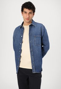 Donkerblauwe denimshirt met een knoopsluiting aan de voorkant en een enkele borstzak. Zachte stof, lange mouwen en een klassiek kraagontwerp.