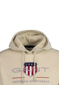 Beige hoodie met een groot geborduurd schildlogo met blauwe, rode en witte accenten. Koordjes en ribgebreide afwerking op de capuchon.