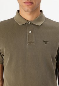 Olivgrünes Polo-Shirt aus strukturiertem Stoff mit einer Knopfleiste mit zwei Knöpfen und gesticktem Logo auf der linken Brust. Kurze Ärmel.