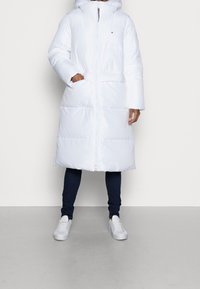 Persona vestida con un abrigo de plumas blanco largo con capucha, pantalones ajustados oscuros y zapatillas blancas, de pie frente a un fondo liso.