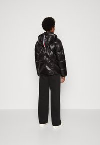 Doudoune noire avec capuche et finition brillante, présentant un matelassage diagonal et un logo de marque à l'arrière, associée à un pantalon à jambes larges.