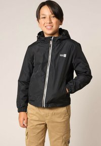 Veste coupe-vent noire avec capuche, fermeture éclair, poignets élastiques, et un patch logo sur la poitrine, portée avec un pantalon cargo kaki.