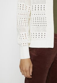 Ruka v krémovém pleteném kardiganu s otevřeným vzorem, odpočívající vedle hnědých kordových kalhot a olivově zelené košile.