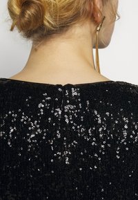 Blondynka z włosami w kok, ubrana w czarną suknię z cekinami i długie złote kolczyki, widziana z tyłu i z boku.