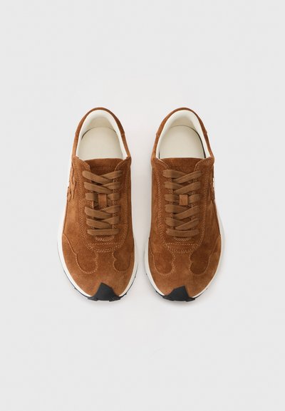 O pereche de adidași din suede maroniu, cu talpă albă și șirete maronii, văzuți de sus pe un fundal deschis.