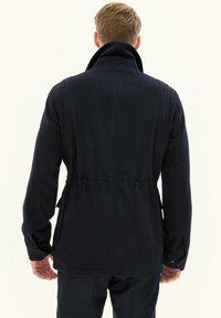 Merinovillainen tummansininen takki, jossa on korkeakauluksinen, säädettävä vyötärö ja pitkät hihat. Takissa on sileä pinta ja huomaamaton sauma yksityiskohtia.