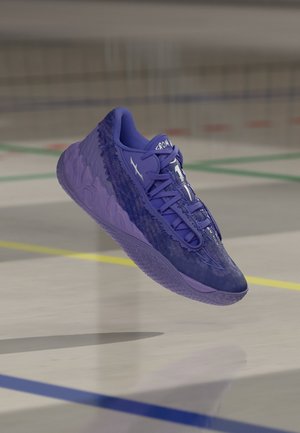 Lilla sportssko med tekstureret sål og detaljeret overdel, svævende over en indendørs sportsbane med farvede afgrænsningslinjer.