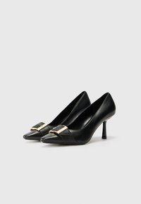 Pantofi cu toc înalt din piele neagră, cu vârfuri ascuțite, dotati cu un accent decorativ din aur pe partea superioară și o textură lucioasă, elegantă.
