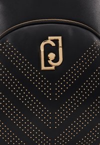 Mochila de imitación de cuero negra con cierre de cremallera, herrajes dorados y un patrón de chevron con tachuelas en el frente. Incluye emblema de logo.