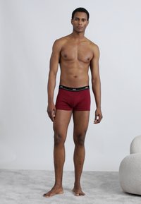 Rode boxershorts met een zwarte elastische tailleband met een logo. Model staat op een lichtgrijs oppervlak tegen een effen achtergrond.