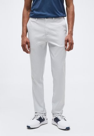 Man draagt lichtgrijze slim-fit broek, witte en marineblauwe sneakers en een donkerblauw shirt tegen een effen achtergrond.