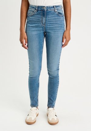 Jeans skinny en denim bleu clair, taille haute avec cinq poches, légère décoloration, portés avec des baskets blanches aux accents beige.