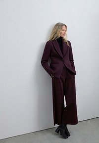 Traje a cuadros burdeos con chaqueta a medida y pantalones de pierna ancha, combinado con un suéter de cuello alto negro y botas negras de tacón.