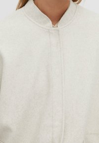 Crème-kleurige vilten jas met een bandkraag, met een strak ontwerp en subtiele textuur, zichtbare stiksels langs de naden van het voorpaneel.