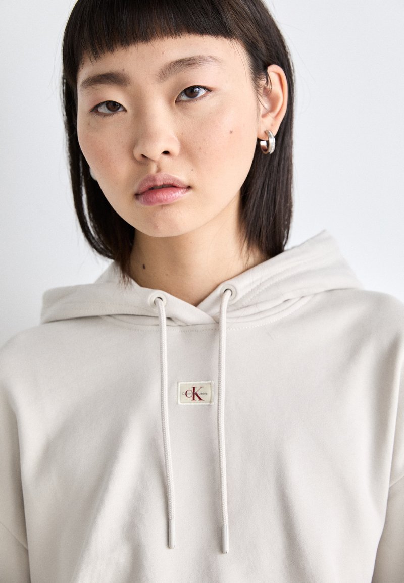 Hellbeige Kapuzenpullover mit Kordelzügen, ausgestattet mit einem kleinen, rechteckigen CK-Logo-Patch. Weicher Stoff mit glatter Textur.