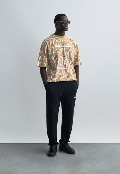 Άνδρας που φοράει πουκάμισο Moschino με ανοιχτό καφέ αφηρημένο σχέδιο, μαύρο παντελόνι τζόγκινγκ, μαύρα αθλητικά παπούτσια και μαύρα γυαλιά ηλίου, στέκεται μπροστά σε ανοιχτό γκρι φόντο.