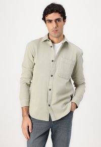 Cămașă butonată de un verde deschis, cu un buzunar pe piept, mâneci lungi și nasturi închiși la culoare, purtată peste un tricou alb. Cumpărată cu blugi gri.
