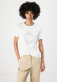 Hvid bomulds t-shirt med et cirkulært grafisk design af en sejlbåd og tekst, parret med beige bukser, der viser en afslappet pasform.