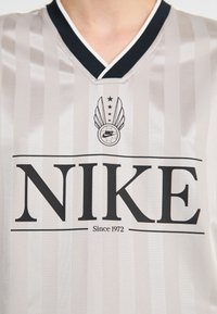 Nike football jersey in light gray with vertical stripes. Features a black v-neck collar, large black logo, and emblem above with wings and stars.