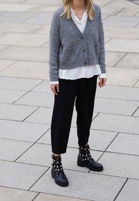 Šedý pletený cardigan s kapsami, položený přes bílou blůzu. Volné černé kalhoty a černé kotníkové boots s perlovými akcenty.