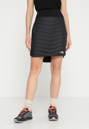 Persona che indossa una gonna trapuntata nera con zip laterali, calzini beige, scarpe da ginnastica nere e rosa e un top nero, in piedi davanti a uno sfondo semplice.