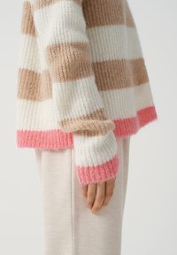 Stickad tröja med breda ränder i kräm, beige och rosa, med en mjuk textur och ribbade ärmslut, bärs över lätta tygbyxor.