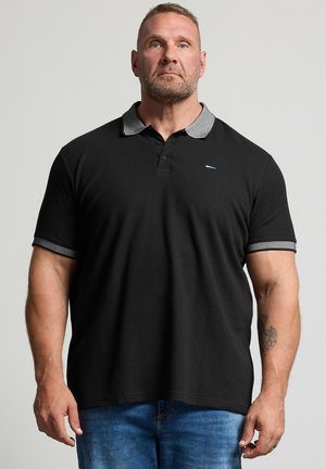 Muscular man with short hair and beard wears black polo shirt with gray collar and blue jeans, standing against plain light background.