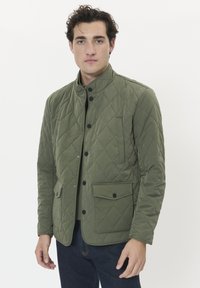 Mann, der eine grüne Steppjacke mit schwarzen Druckknöpfen trägt, steht vor einem weißen Hintergrund, die Hände entspannt an den Seiten.