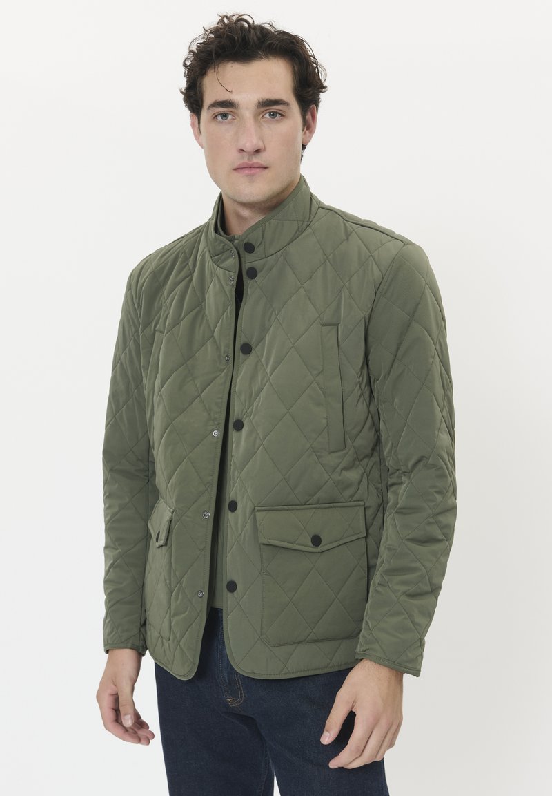 Mann, der eine grüne Steppjacke mit schwarzen Druckknöpfen trägt, steht vor einem weißen Hintergrund, die Hände entspannt an den Seiten.