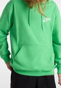 Jasnozielona bluza Vans z kieszenią kangurzą z przodu oraz logo na lewej piersi, noszona z czarnymi spodniami, częściowy widok na osobę.