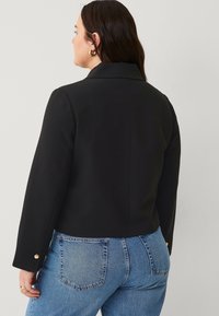 Black cropped jacket with a tailored collar, long sleeves, and gold button accents on cuffs, paired with classic blue jeans.