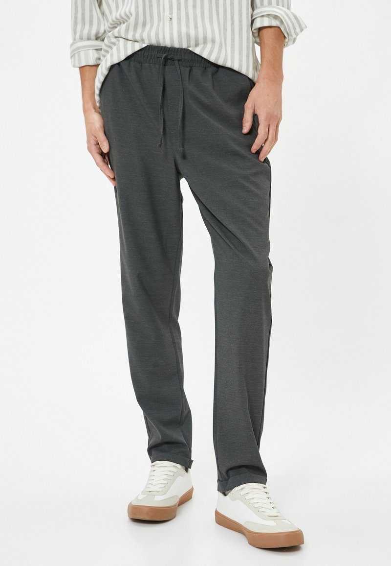 Grå bukser med elastisk talje og snor. Lige snit, blød tekstur. Sammen med hvide og beige sneakers, på en ensfarvet baggrund.