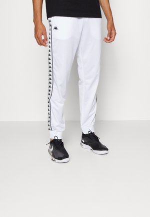 Homme portant un pantalon de survêtement blanc avec une bande noire à logo sur les côtés et des baskets noires, sur un fond uni.