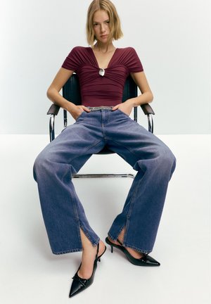 Femeie blondă stând pe un scaun negru, purtând un top marsat bordo cu o broșă argintie, blugi albaștri evazați cu fante în față și pantofi negri cu toc ascuțit.