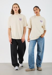 Beige bomull t-skjorter med lilla grafikk, løs passform. Modellen til venstre har på seg svarte vide bukser, modellen til høyre har lys blå jeans og beige sko.