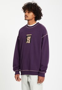Paarse sweatshirt van zachte stof, met een grafisch ontwerp aan de voorkant met zwarte en beige accenten, en contrasterende stiksels.