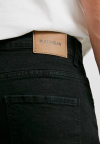 Gros plan sur la poche arrière d'un jean noir avec un patch en cuir beige portant l'inscription "PULL&BEAR", porté avec une chemise blanche.