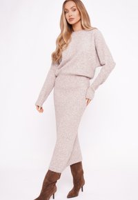 Kobieta ubrana w beżowy dziany sweter i pasującą długą spódnicę oraz brązowe zamszowe botki na wysokim obcasie, pozująca na tle białego tła.