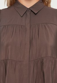 Brown blouse featuring a collared neck, button closure, and gathered details at the chest and waist, made from smooth, lightweight fabric.