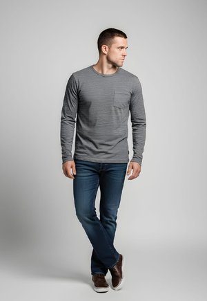 Hombre de pie con las piernas cruzadas, vestido con una camisa gris de manga larga con bolsillo, vaqueros azules y zapatos casuales marrones, mirando hacia un lado.