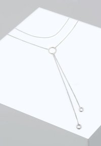 Sølv metal halskæde med en cirkulær vedhæng, hvor to kæder strækker sig nedad, hver ender i en mindre cirkel. Glat overflade, minimalistisk design.