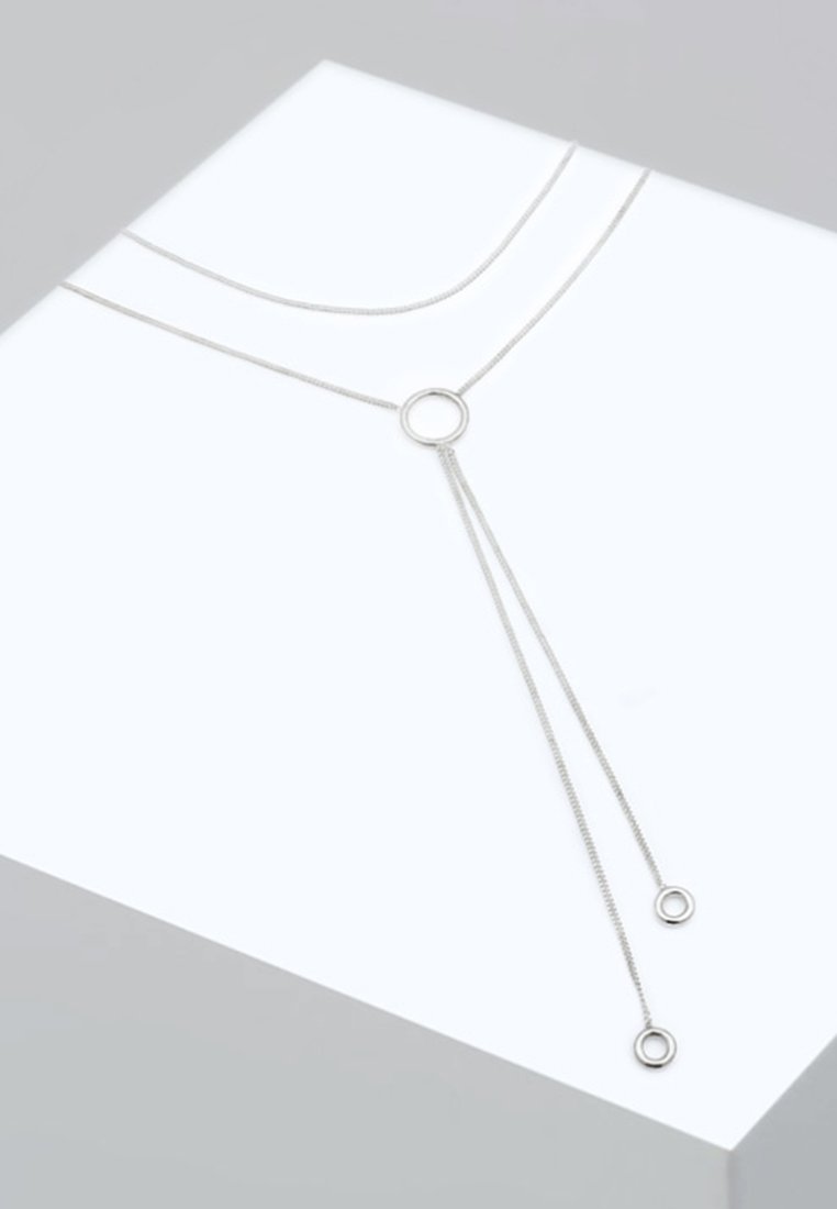 Sølv metal halskæde med en cirkulær vedhæng, hvor to kæder strækker sig nedad, hver ender i en mindre cirkel. Glat overflade, minimalistisk design.