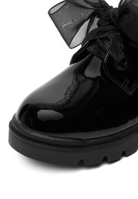 Bota de tobillo de charol negro con punta redonda, suela de goma texturizada y un lazo negro translúcido en los cordones.