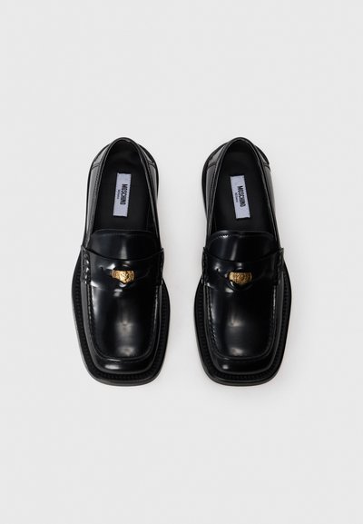 Ein Paar glänzende schwarze Moschino Lederloafers mit quadratischen Zehen und goldenem Emblem auf weißem Hintergrund.