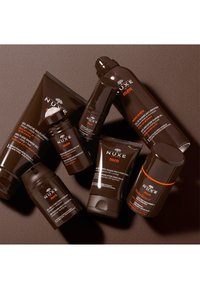 En samling av NUXE Men groomingprodukter i mörkbruna tuber och flaskor, med vit och orange text, i olika former och storlekar.