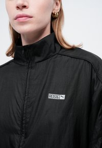 Kvinde iført sort zip-up jakke med høj krave og gyldne ringsøreringe, der viser "ROSIE" og Puma-logoet på brystet.