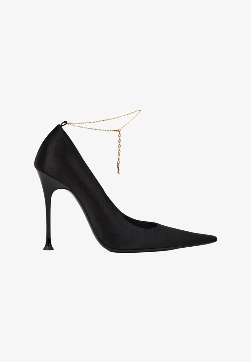 Chaussure noire à talon haut, bout pointu, talon aiguille fin et fine bride de cheville en chaîne dorée sur fond blanc.