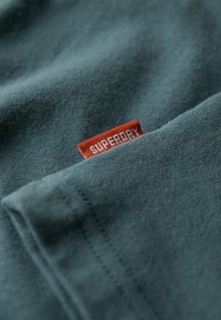 Tejido de algodón en color verde azulado con una textura suave, que presenta una etiqueta roja con "SUPERDRY" en blanco. Enfoque en los detalles de costura en los bordes.
