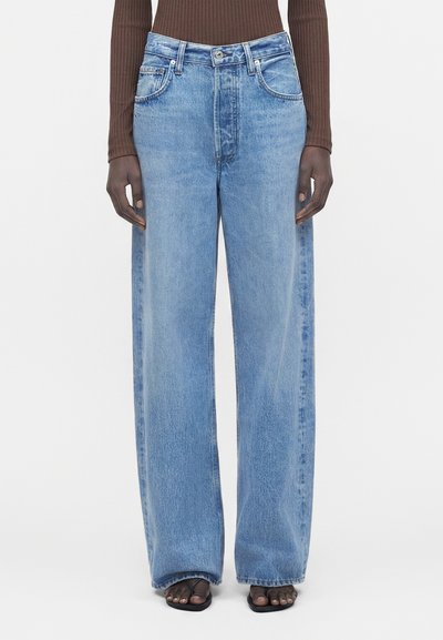 Model wearing light blue wide-leg jeans, brown ribbed long sleeve top, and black open-toe sandals against white background.